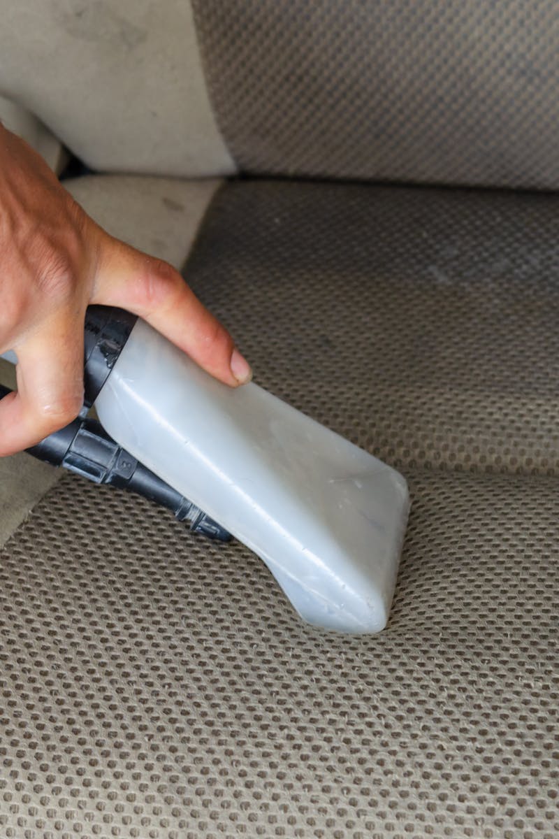 Hands using a vacuum nozzle to deeply clean a fabric car seat, removing dirt and stains.